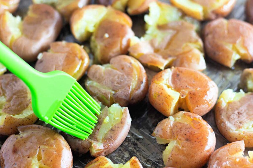 A green silicone brush spreads oil over smashed roasted potatoes on a baking tray.