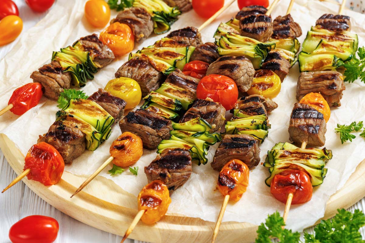 Grilled skewers with beef, zucchini slices, cherry tomatoes, and herbs arranged on a wooden board with parchment paper.