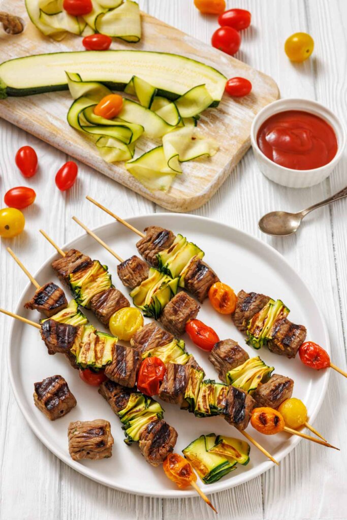 Grilled beef, zucchini ribbons, and cherry tomatoes on skewers arranged on a white plate, with a cutting board of sliced zucchini and a bowl of sauce beside it.