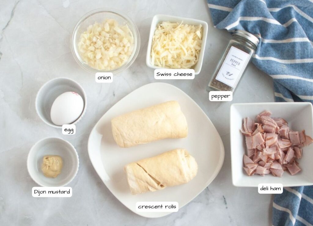 Overhead shot of labeled ingredients for Ham and Cheese Puffs, including crescent rolls, deli ham, Swiss cheese, onion, egg, Dijon mustard, and pepper.