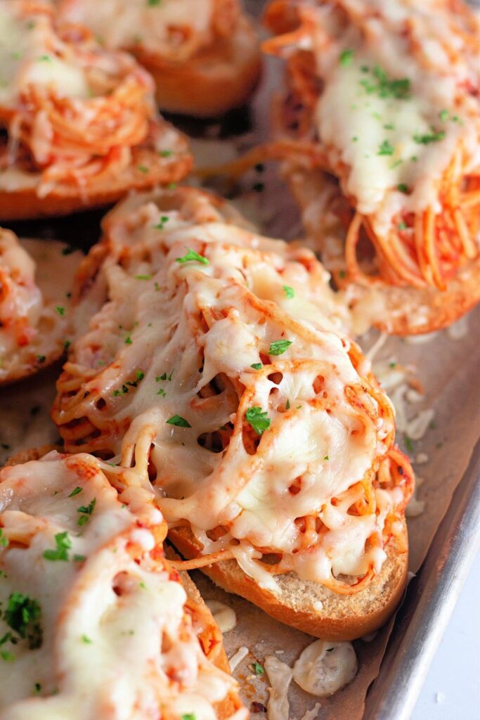 Slices of bread topped with spaghetti, tomato sauce, melted cheese, and sprinkled with chopped parsley on a baking sheet.