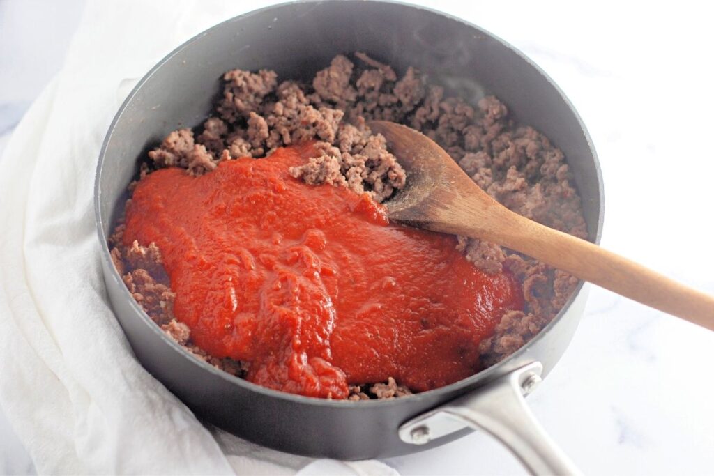 Ground beef and tomato sauce in a black skillet being stirred with a wooden spoon, set on a white surface with a white cloth underneath.