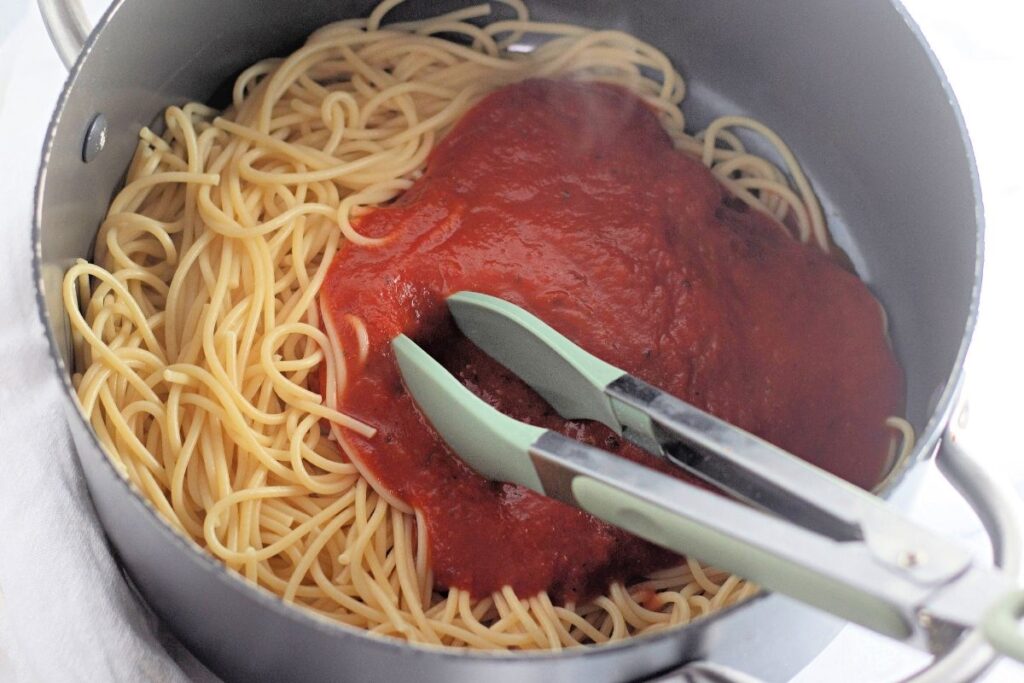Cooked spaghetti with tomato sauce and metal tongs in a large pot on a white surface.