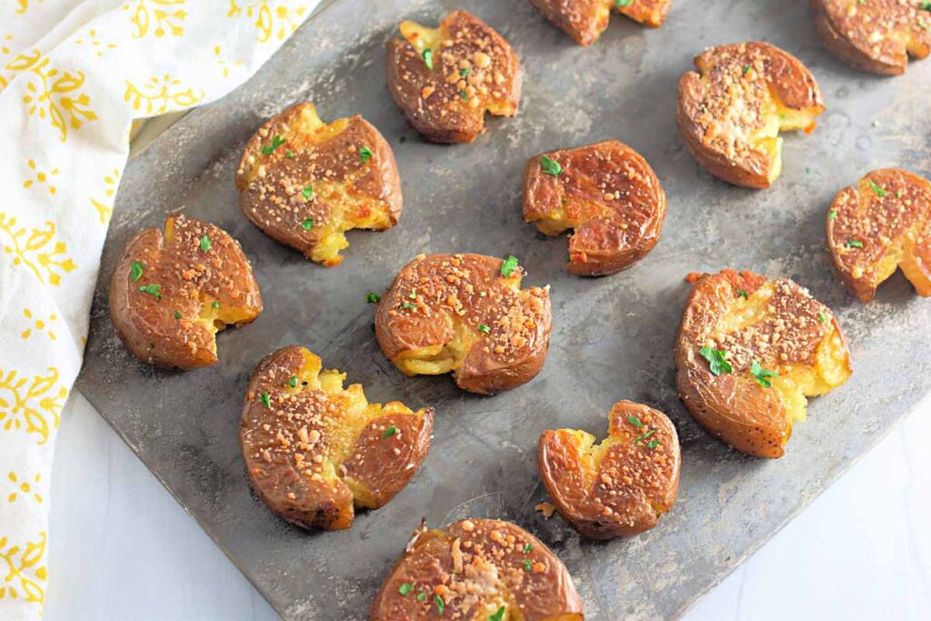 Baked smashed potatoes topped with seasoning and herbs arranged on a metal baking sheet.