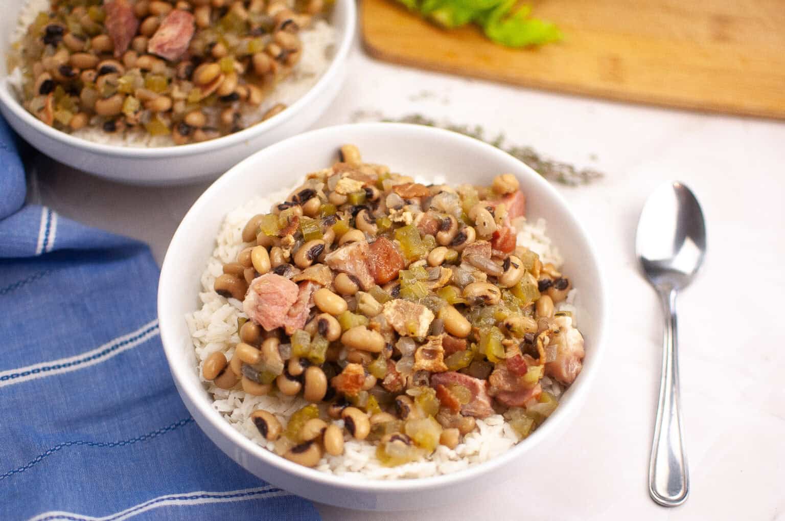 A bowl of Hoppin&rsquo; John served over white rice, with a spoon and a blue napkin nearby.