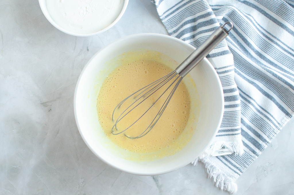 Overhead shot of a pale yellow lemon curd mixture being whisked in a white bowl.