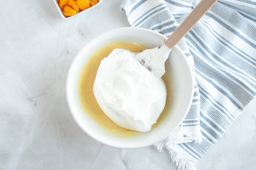 Overhead shot of white whipped topping being gently folded into a pale yellow mixture in a white bowl with a spatula.