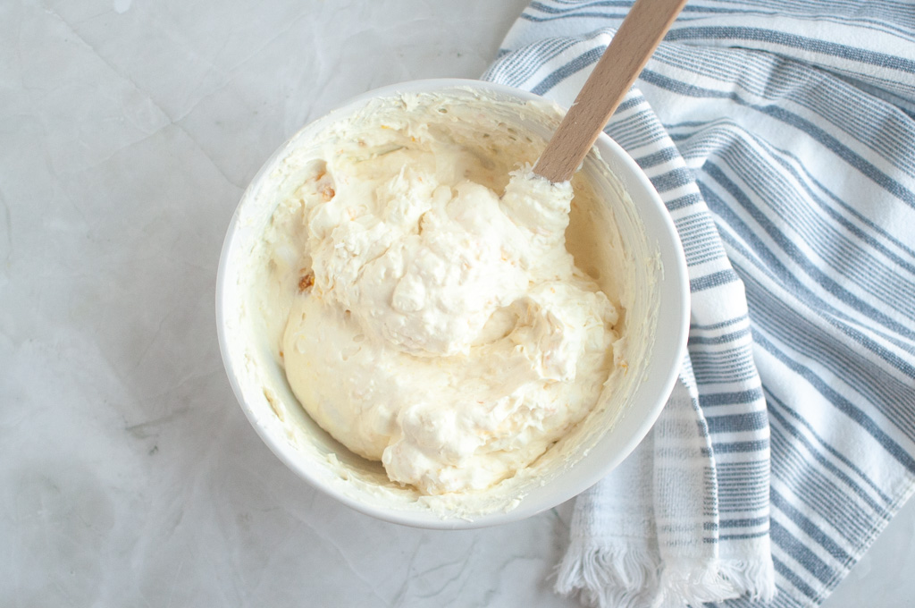 Overhead shot of a white bowl filled with creamy, light-colored Orange Fluff Salad being mixed with a spatula.