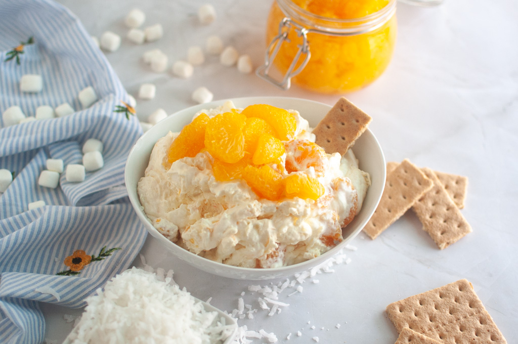 Overhead shot of a white bowl filled with creamy Orange Fluff Salad, topped with mandarin oranges and served with a graham cracker.