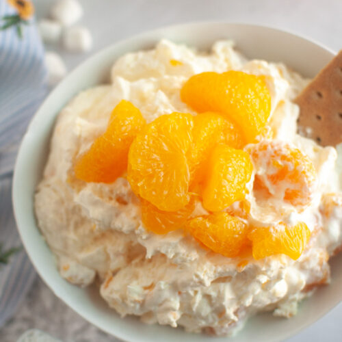 Overhead shot of a white bowl filled with creamy Orange Fluff Salad, topped with mandarin oranges and served with a graham cracker.