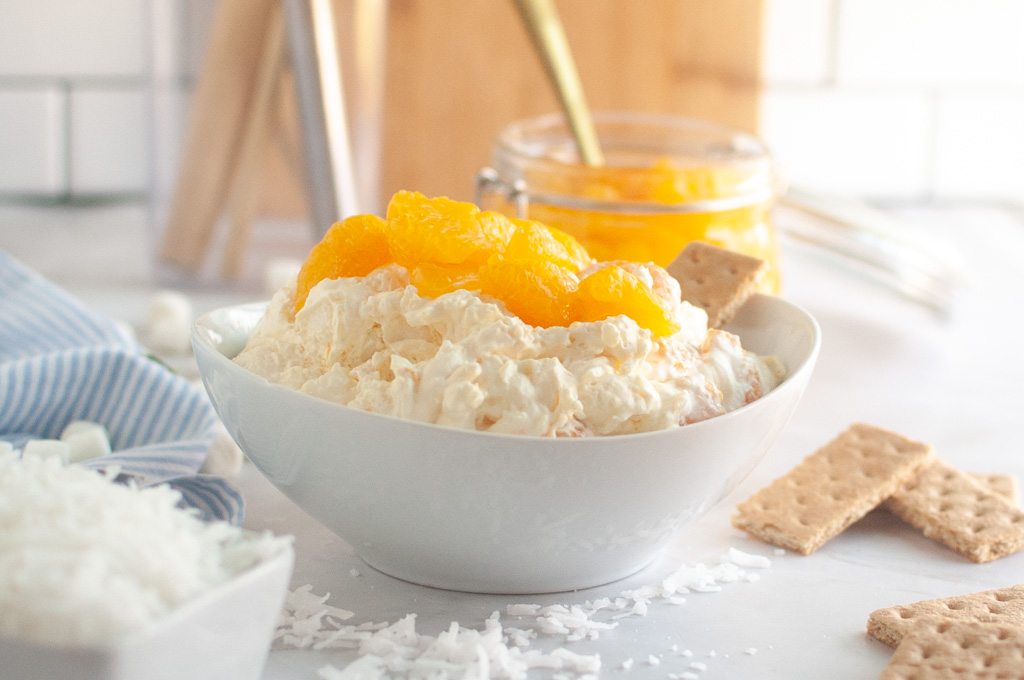 Overhead shot of a white bowl filled with creamy Orange Fluff Salad, topped with mandarin oranges and served with graham crackers.