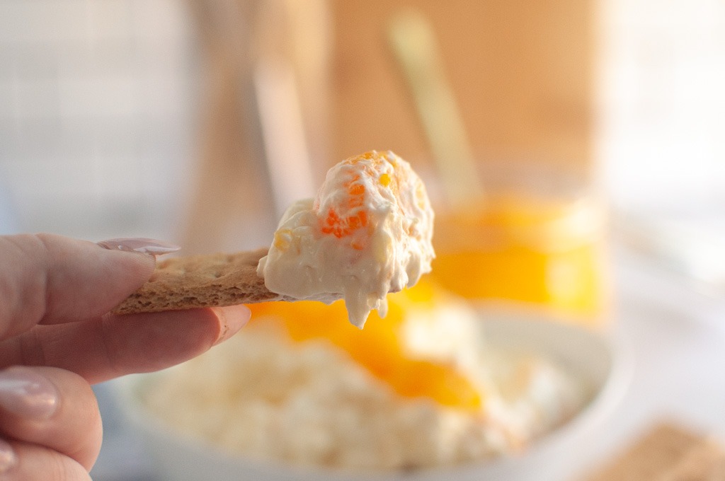 A hand holds a graham cracker topped with creamy Orange Fluff Salad Dip over a blurred bowl of the dip.