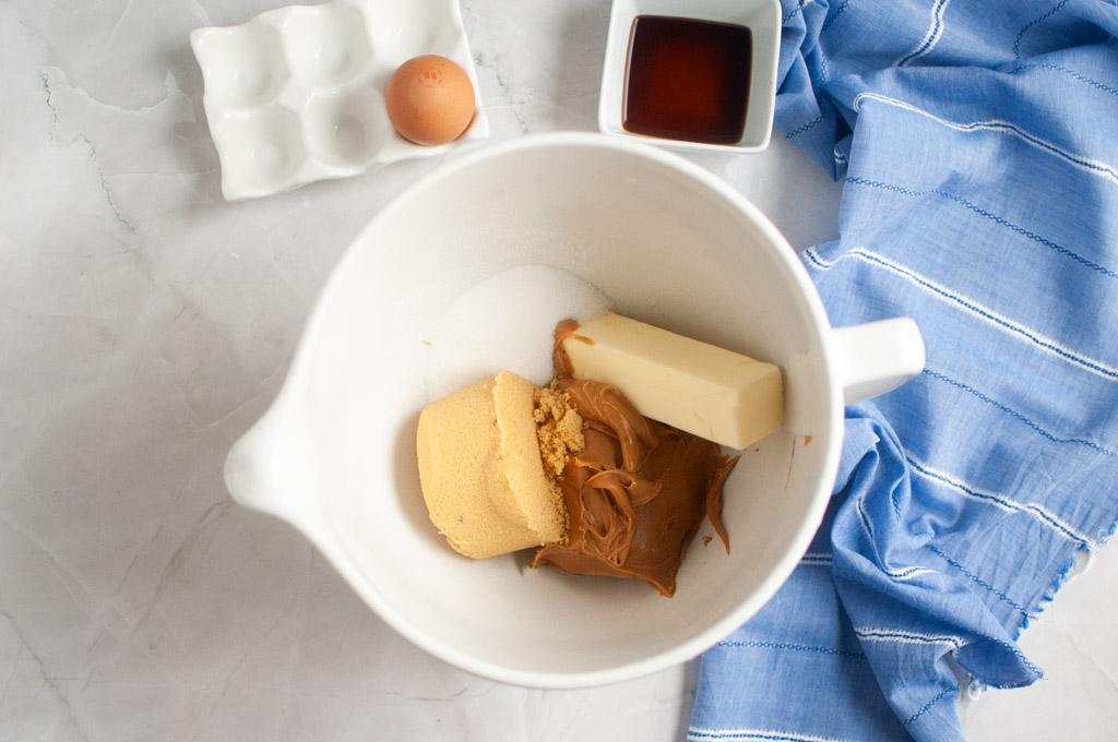 Overhead shot of a white mixing bowl containing peanut butter, butter, and brown sugar, with other ingredients nearby.