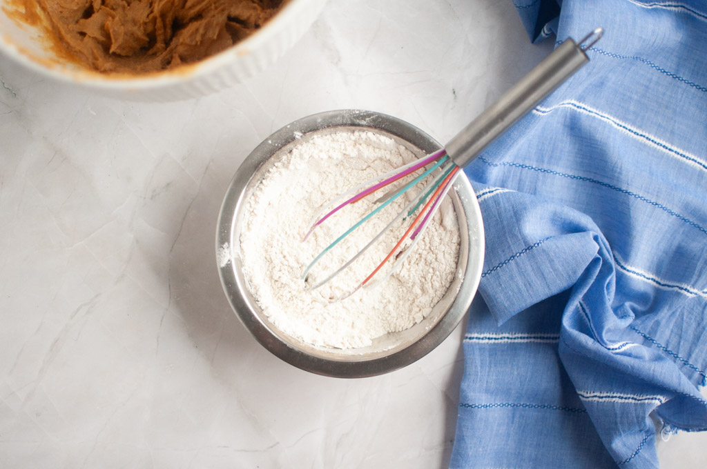 Overhead shot of a metal bowl containing white flour with a whisk resting inside, next to a bowl of peanut butter cookie dough.