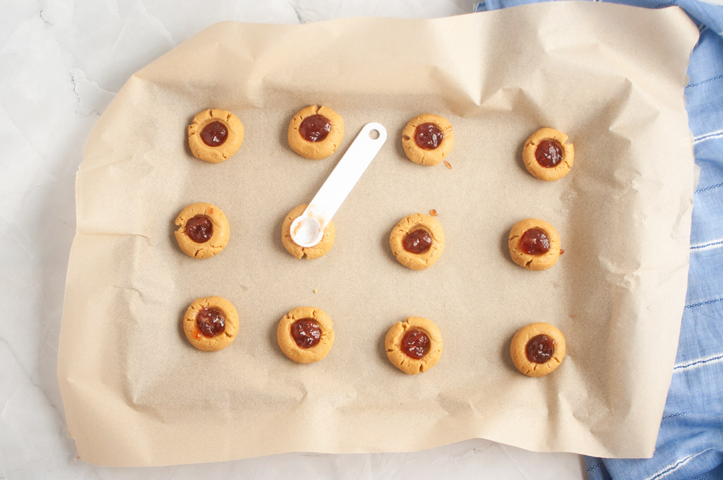 Overhead shot of a baking sheet with peanut butter thumbprint cookies, some being filled with red jelly using a white spoon.