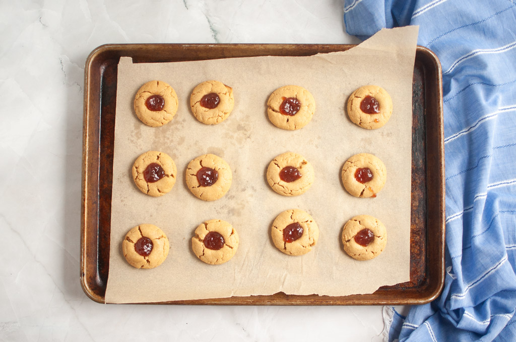 Overhead shot of a baking sheet with twelve peanut butter thumbprint cookies filled with red jelly.