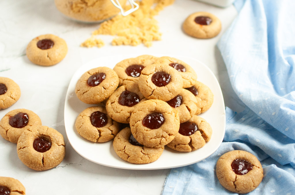A white plate piled with golden-brown peanut butter cookies with a red jelly center, surrounded by more cookies.