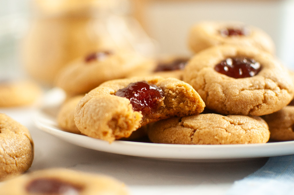 A close-up of a partially eaten peanut butter cookie with a red jelly filling on a white plate.