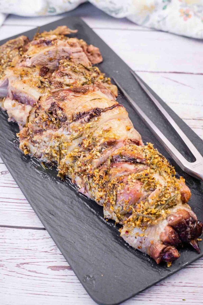 A black platter with slices of roasted leg of lamb, heavily seasoned with herbs, next to a carving fork.