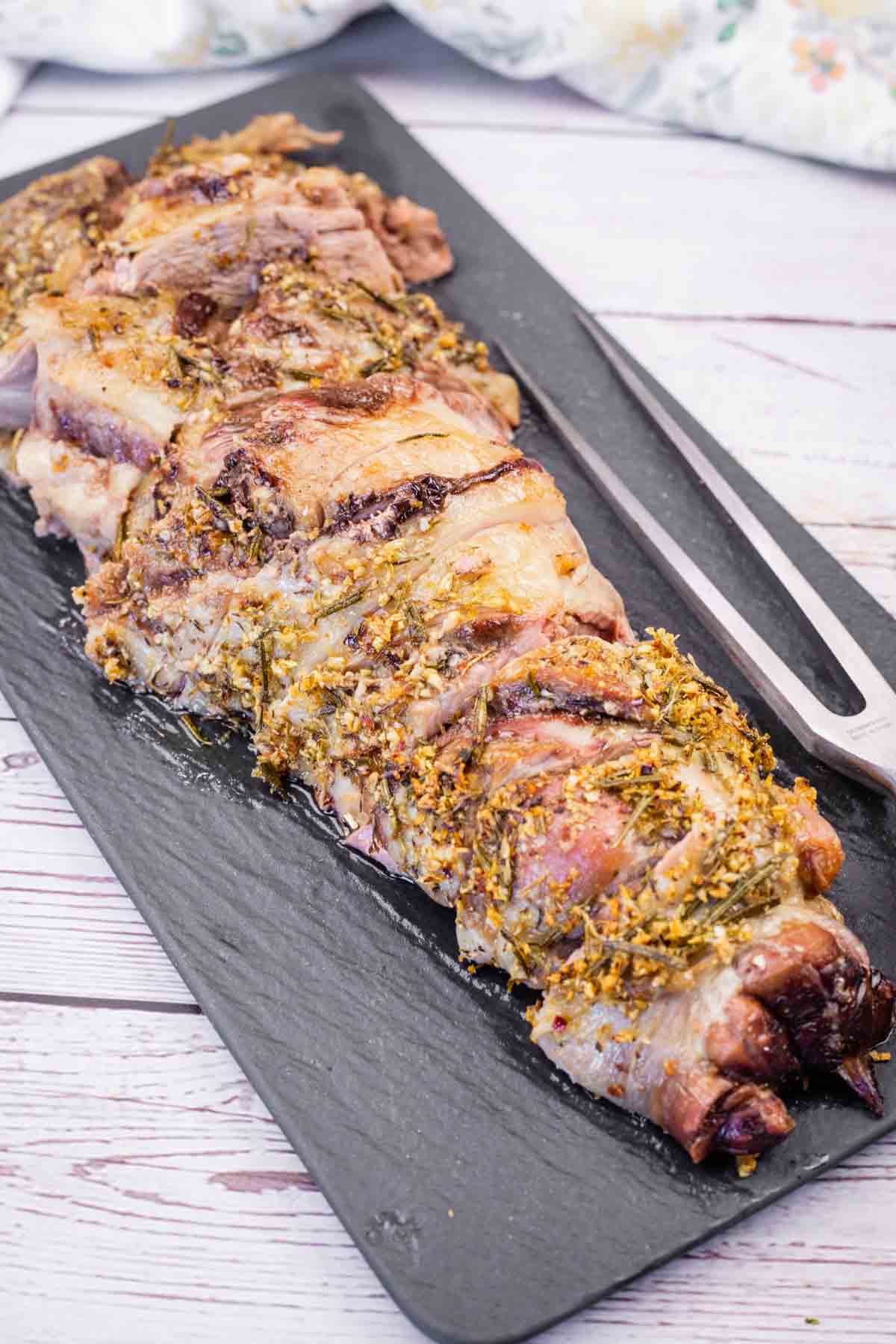 A black platter with slices of roasted leg of lamb, heavily seasoned with herbs, next to a carving fork.
