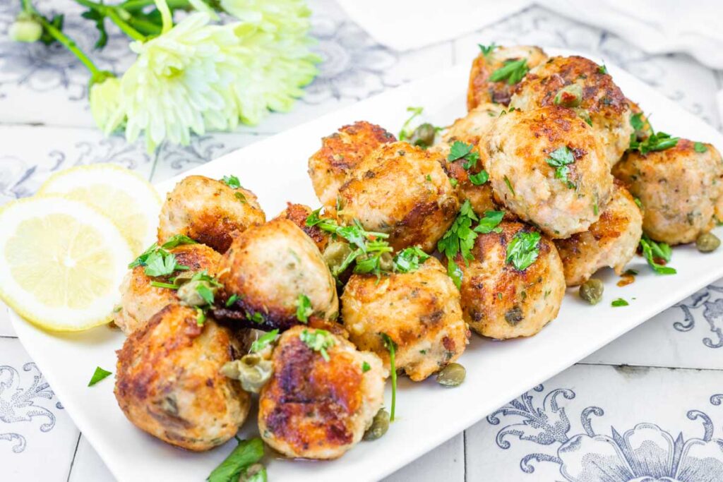 A pile of golden-brown salmon balls garnished with fresh parsley and capers, served with lemon slices on a white plate.
