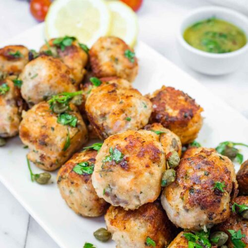 A white plate piled with golden-brown salmon balls garnished with parsley and capers, served with lemon slices and a small bowl of green sauce.