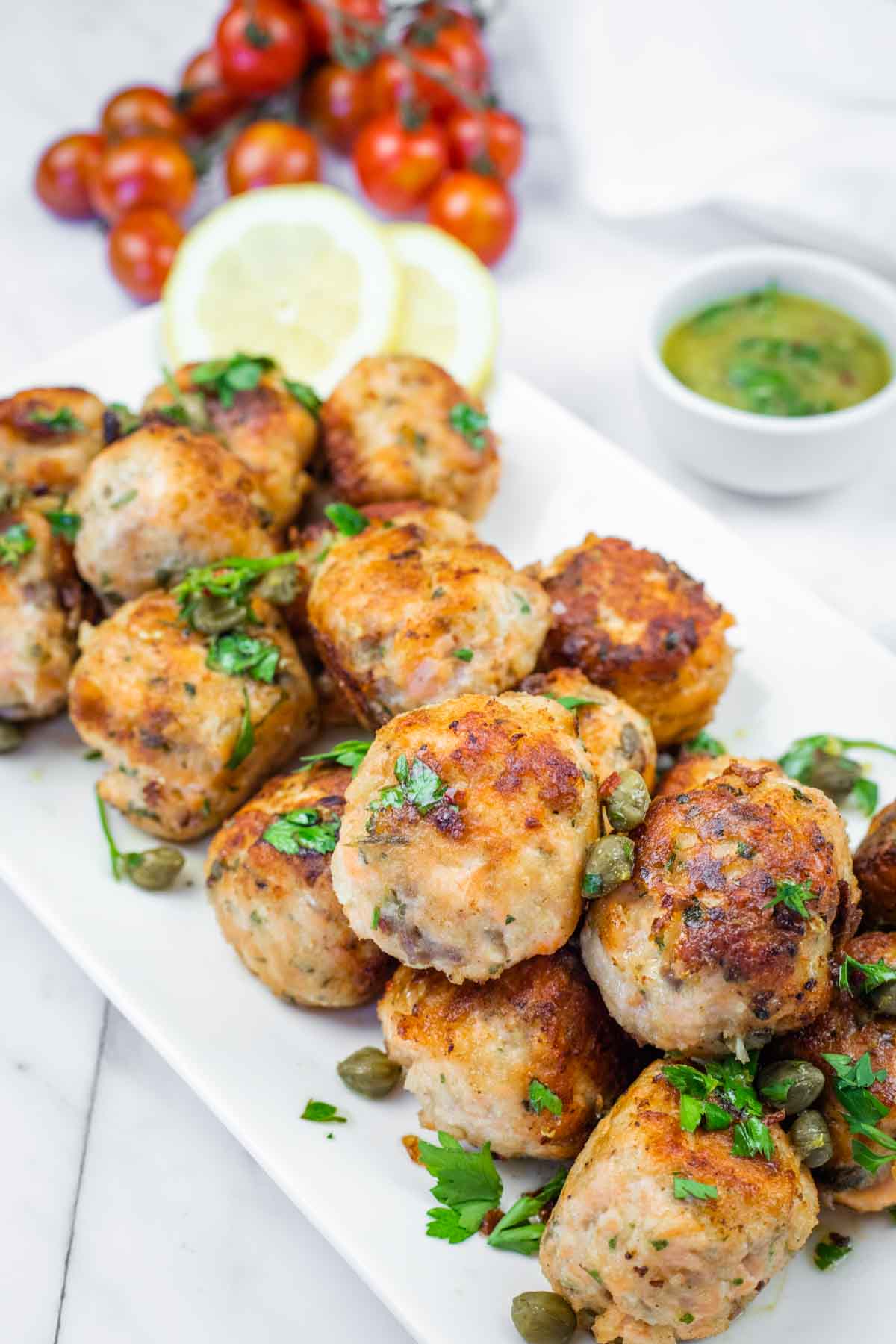 A white plate piled with golden-brown salmon balls garnished with parsley and capers, served with lemon slices and a small bowl of green sauce.