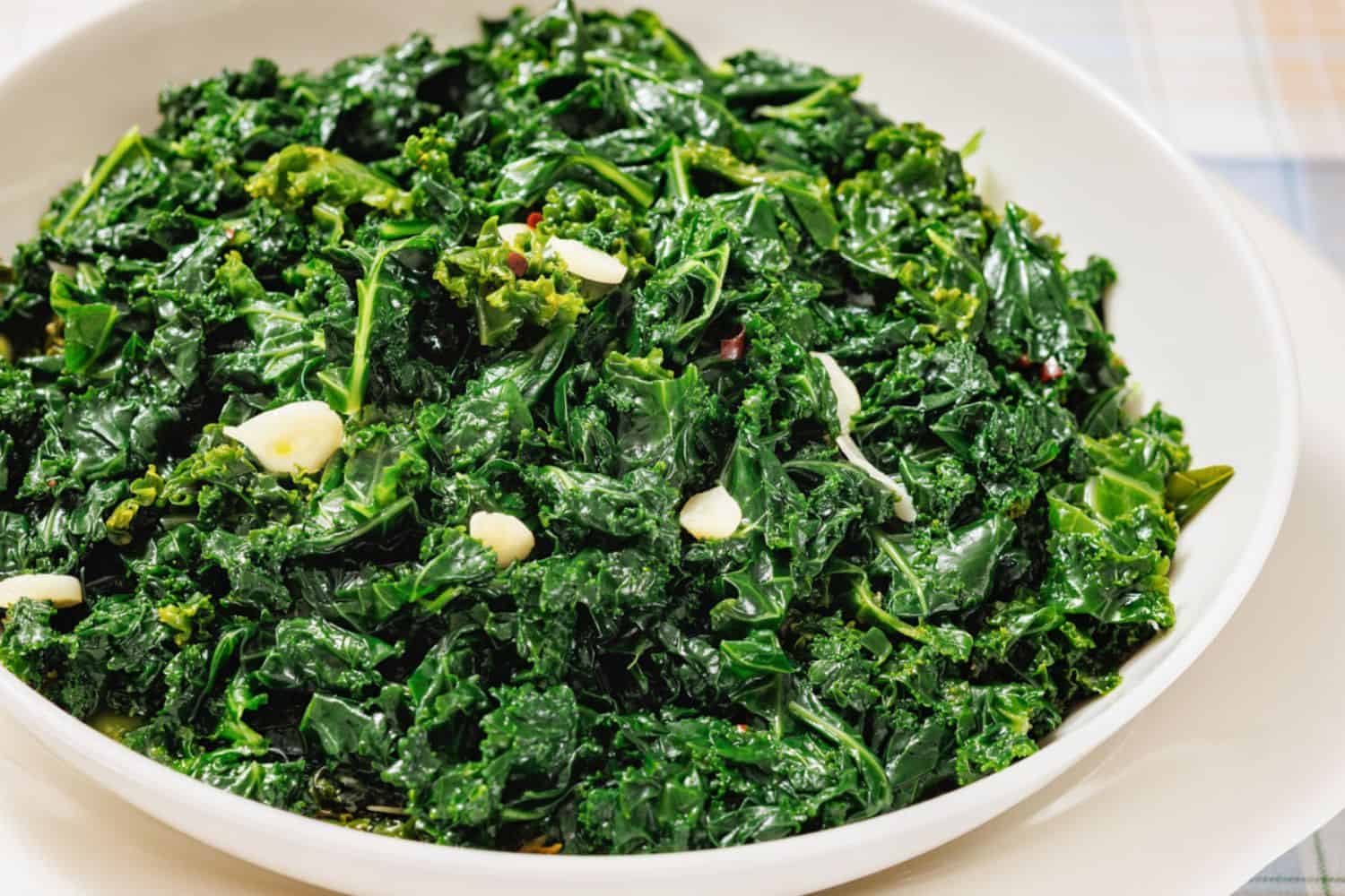 A white bowl filled with saut&eacute;ed kale and sliced garlic on a light-colored surface.