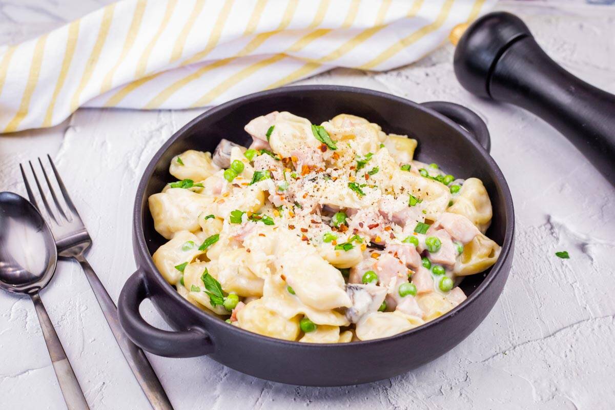 A black bowl of Easy Skillet Cheese Tortellini with Ham & Peas garnished with grated cheese and herbs.