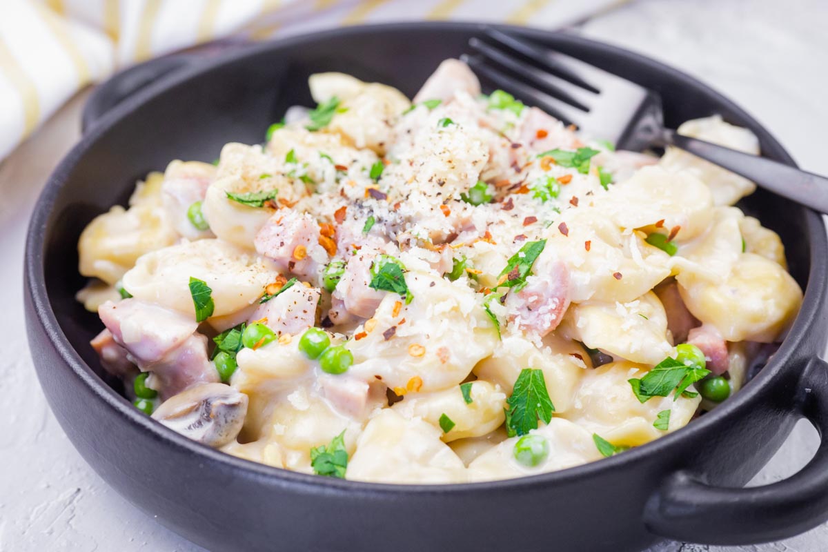 A black bowl filled with Skillet Cheese Tortellini with Ham & Peas, topped with grated cheese and red pepper flakes, with a fork resting on the side.