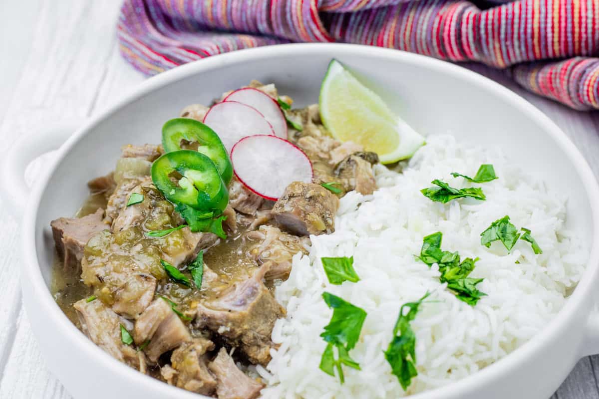 A bowl of white rice and green chili verde garnished with cilantro, radish slices, jalape&ntilde;o, and a lime wedge, set on a white surface with a colorful cloth in the background.