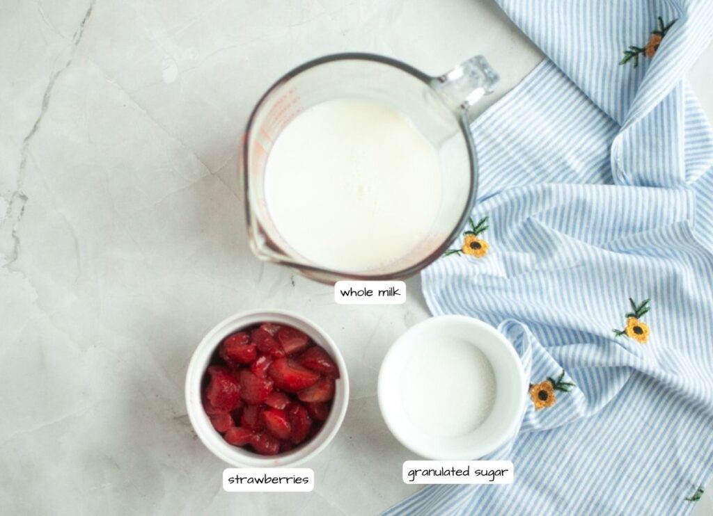 Overhead shot of sliced strawberries, whole milk, and granulated sugar on a marble surface with a blue and white cloth.