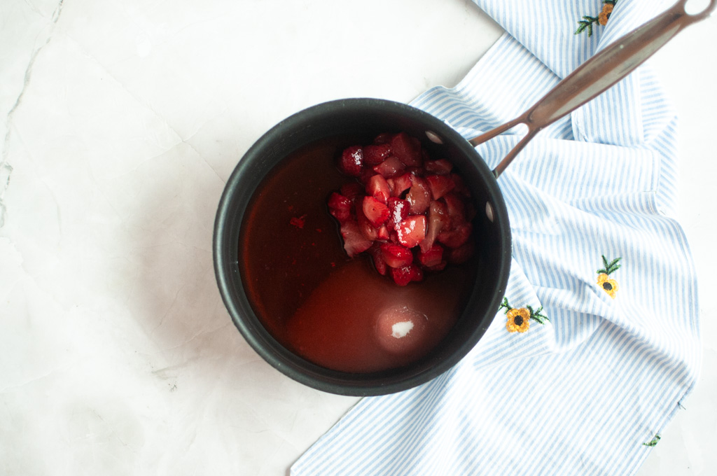 Overhead shot of sliced strawberries in a saucepan with a red liquid, likely being cooked into a sauce or compote.