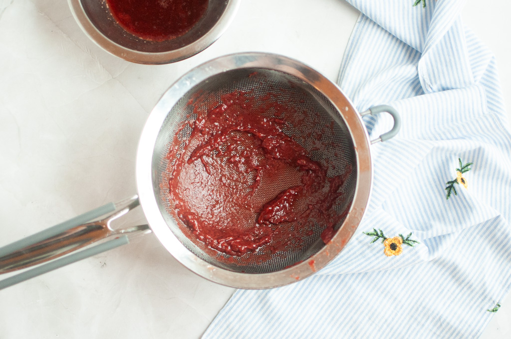 Overhead shot of a metal strainer containing thick, dark red red velvet cake batter.