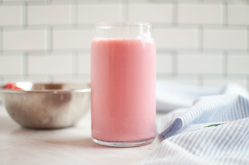 A glass filled with a pink strawberry milk on a white surface with a blue and white cloth.