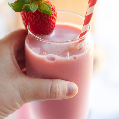 A hand holding a clear glass of pink strawberry milk garnished with a fresh strawberry and a red and white straw.