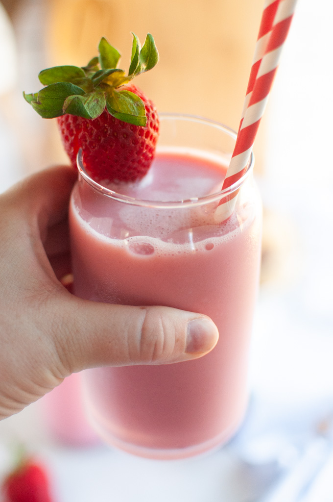 A hand holding a clear glass of pink strawberry milk garnished with a fresh strawberry and a red and white straw.