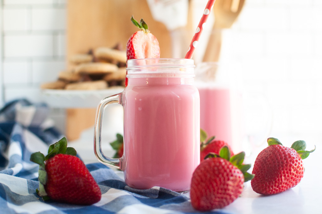 Two pink strawberry milk in glass jars with straws and strawberry garnishes, on a blue and white checkered cloth.