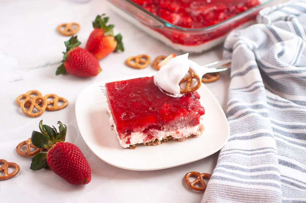 A slice of Strawberry Pretzel Salad topped with whipped cream and a pretzel sits on a white plate.