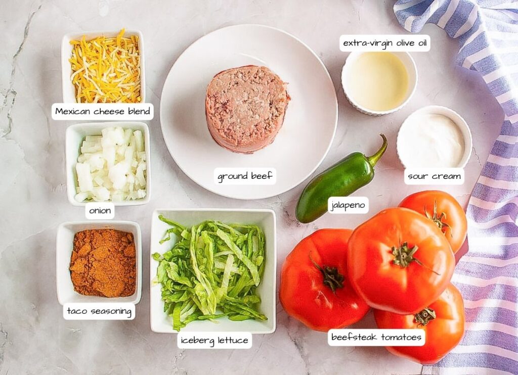 Overhead shot of labeled ingredients for taco stuffed tomatoes, including ground beef, tomatoes, cheese, onion, lettuce, taco seasoning, jalapeño, olive oil, and sour cream.