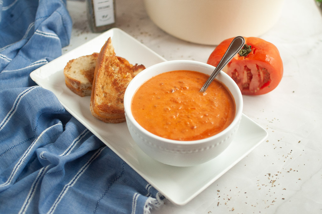 A bowl of tomato soup with a spoon, served with two slices of toasted bread on a white plate; a halved tomato and a blue cloth are nearby.