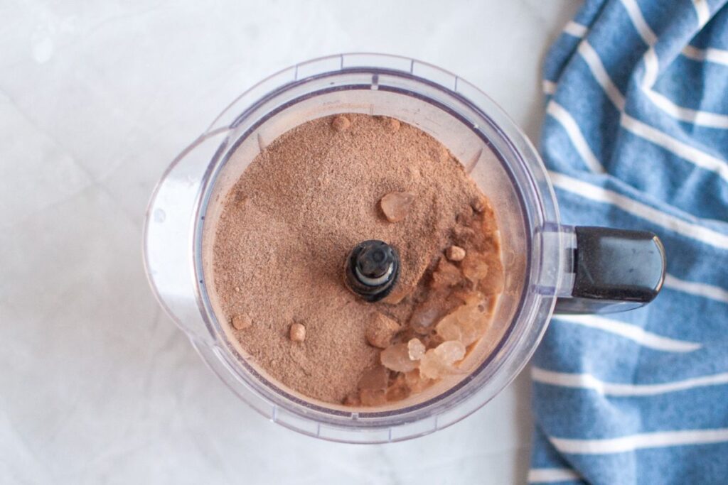 Overhead view of a clear food processor bowl containing a brown hot chocolate mix powder and several pieces of crushed ice, on a white surface with a blue and white striped napkin.