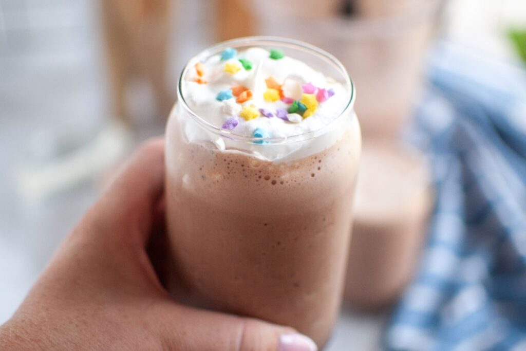 A hand with light pink fingernails holds a clear glass filled with frothy brown frozen hot chocolate, topped with white whipped cream and colorful star sprinkles.