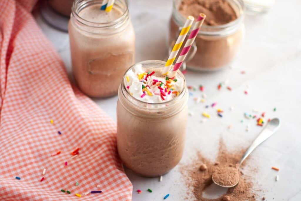 A mason jar filled with a Frozen Hot Chocolate topped with whipped cream and sprinkles, two colorful straws, surrounded by jars of cocoa powder and a pink checkered cloth.
