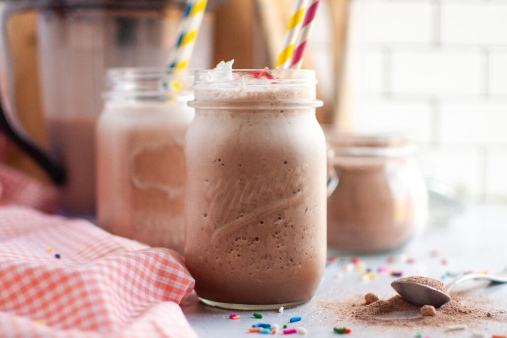 Three clear mason jars filled with frothy brown frozen hot chocolate, topped with whipped cream, sprinkles, and colorful striped straws, with a spoon of cocoa powder on the table.