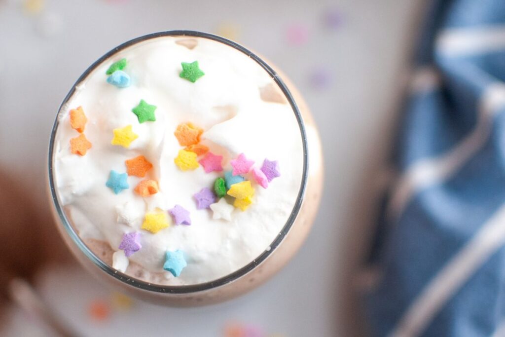 A close-up, top-down shot of a clear glass filled with white whipped cream, generously topped with colorful star-shaped sprinkles, covering a light brown frozen hot chocolate.