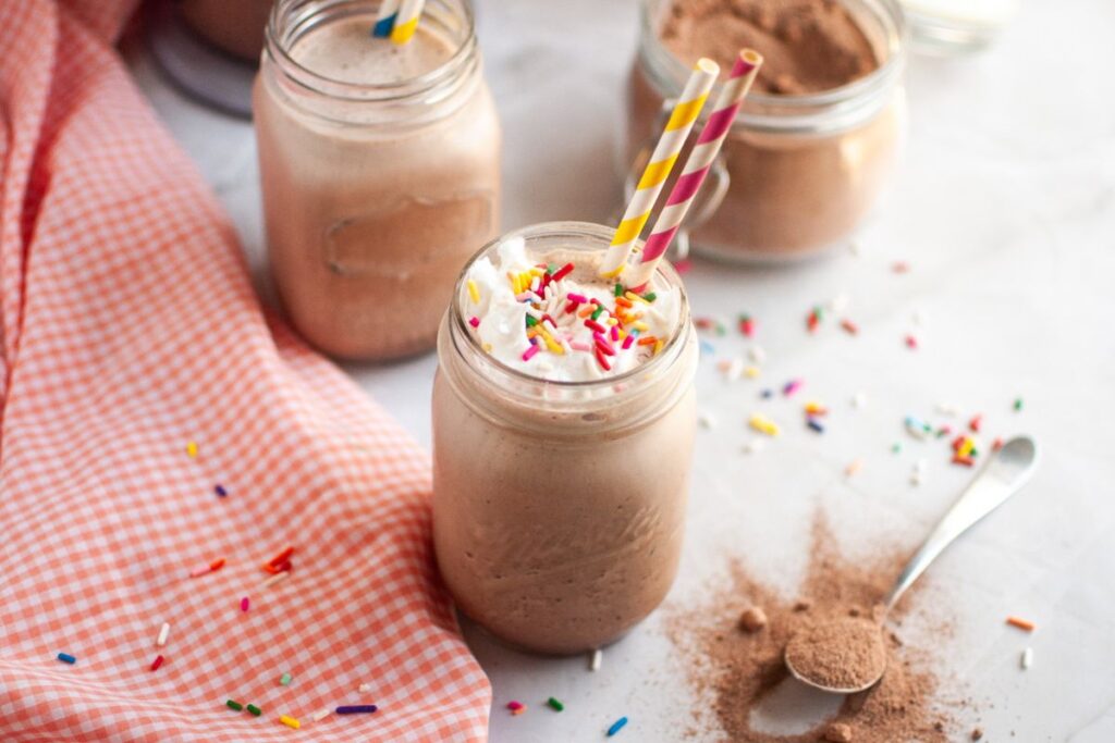 Two clear mason jars filled with frothy brown frozen hot chocolate, topped with whipped cream, sprinkles, and colorful striped straws, with a jar of cocoa mix and cocoa powder on the table.