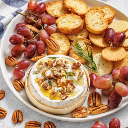 A slightly high-angle shot of a white circular platter featuring a baked round of brie cheese drizzled with honey, topped with chopped pecans and a sprig of fresh rosemary. The platter is surrounded by bunches of red grapes and an assortment of golden-brown crackers and crostini.