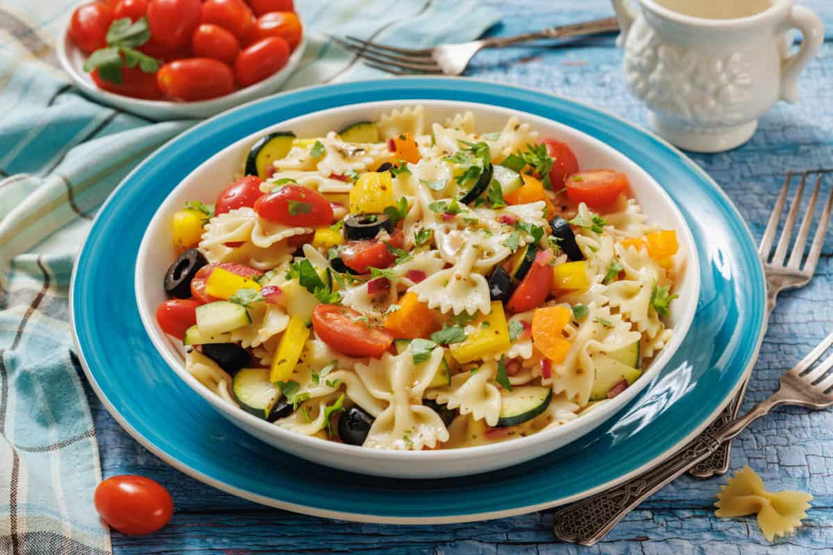 A plate of bowtie pasta salad with cherry tomatoes, yellow bell peppers, zucchini, black olives, and herbs, set on a blue plate with a bowl of tomatoes and utensils nearby.