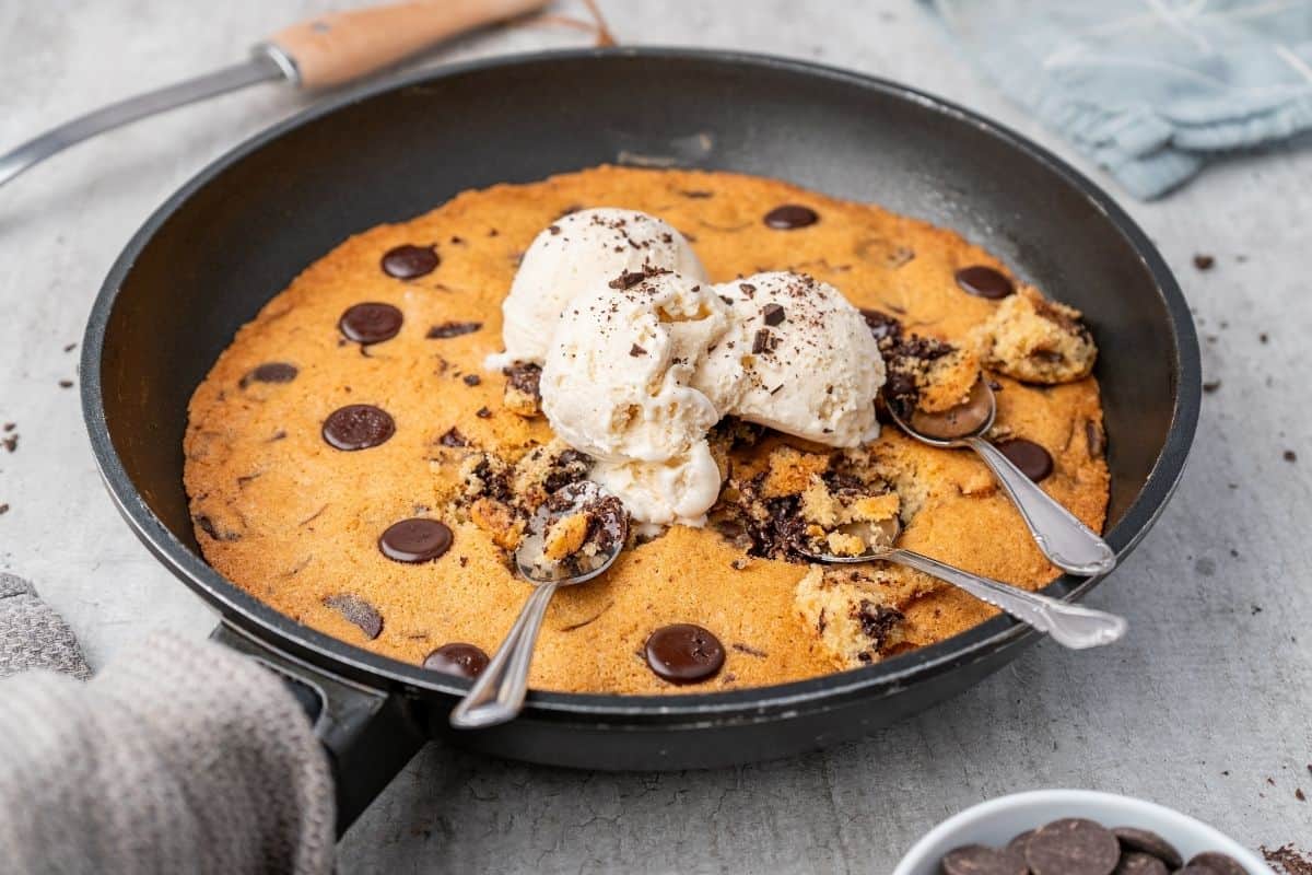 Overhead shot of a warm chocolate chip skillet cookie in a black skillet, topped with scoops of vanilla ice cream and three spoons.